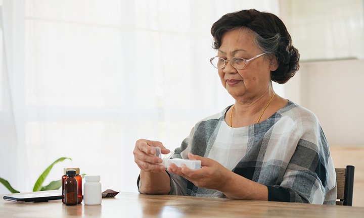 Mature Asian woman in black and white sweater wearing glasses takes daily medication while sitting down.